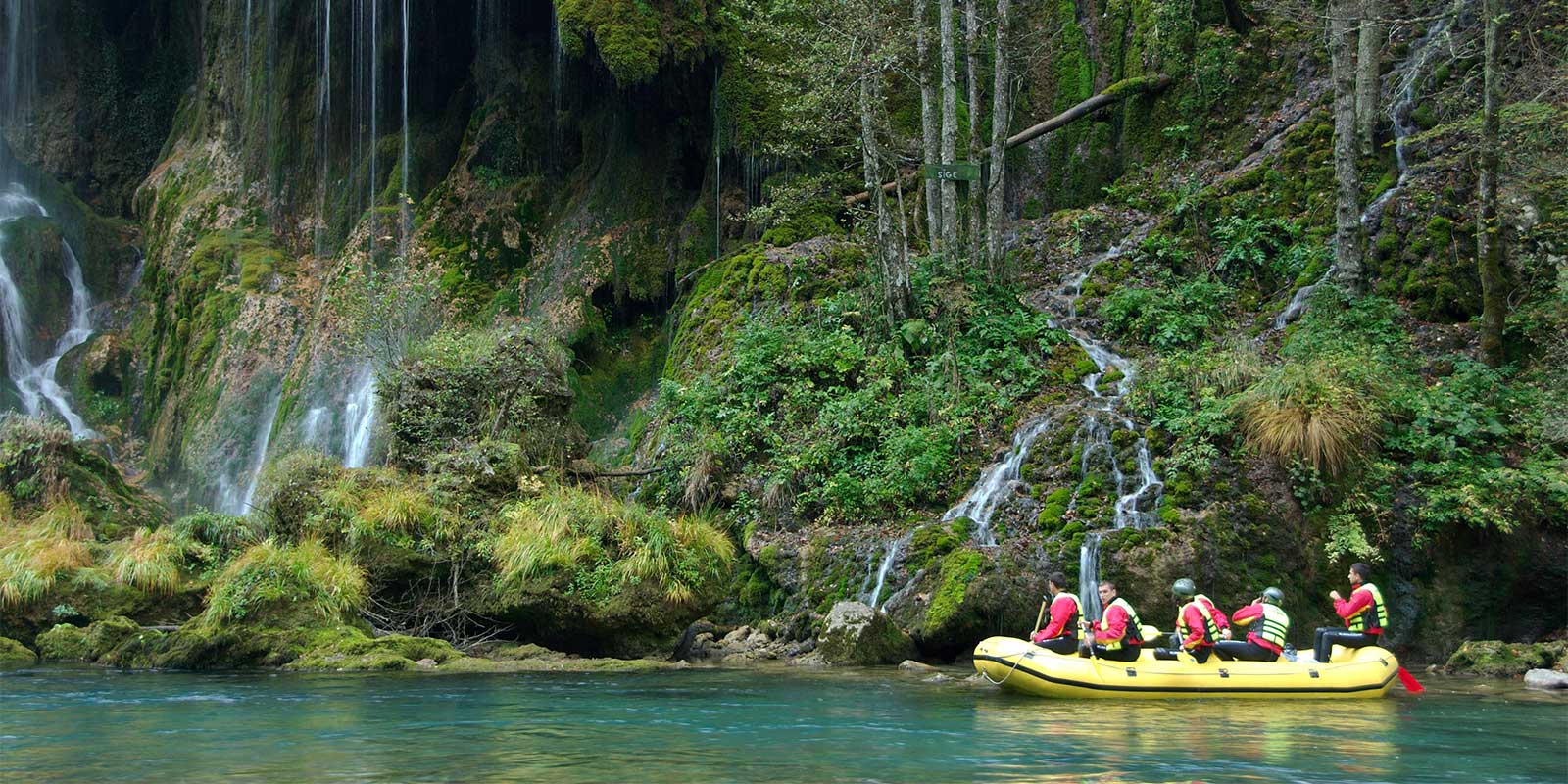 Group in inflatable raft on river with waterfall coming down cliff behind